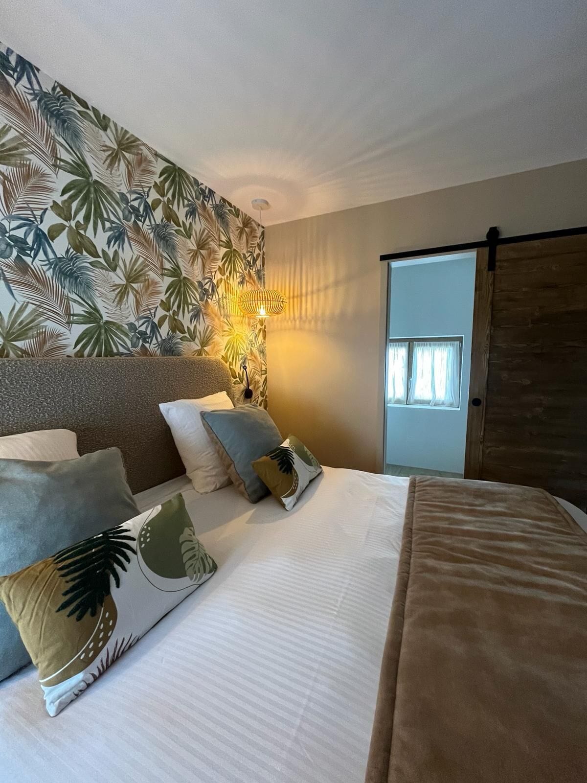 Een slaapkamer met een bed, kussens, een muur met een tropisch bladpatroon, een hanglamp en een schuifdeur.