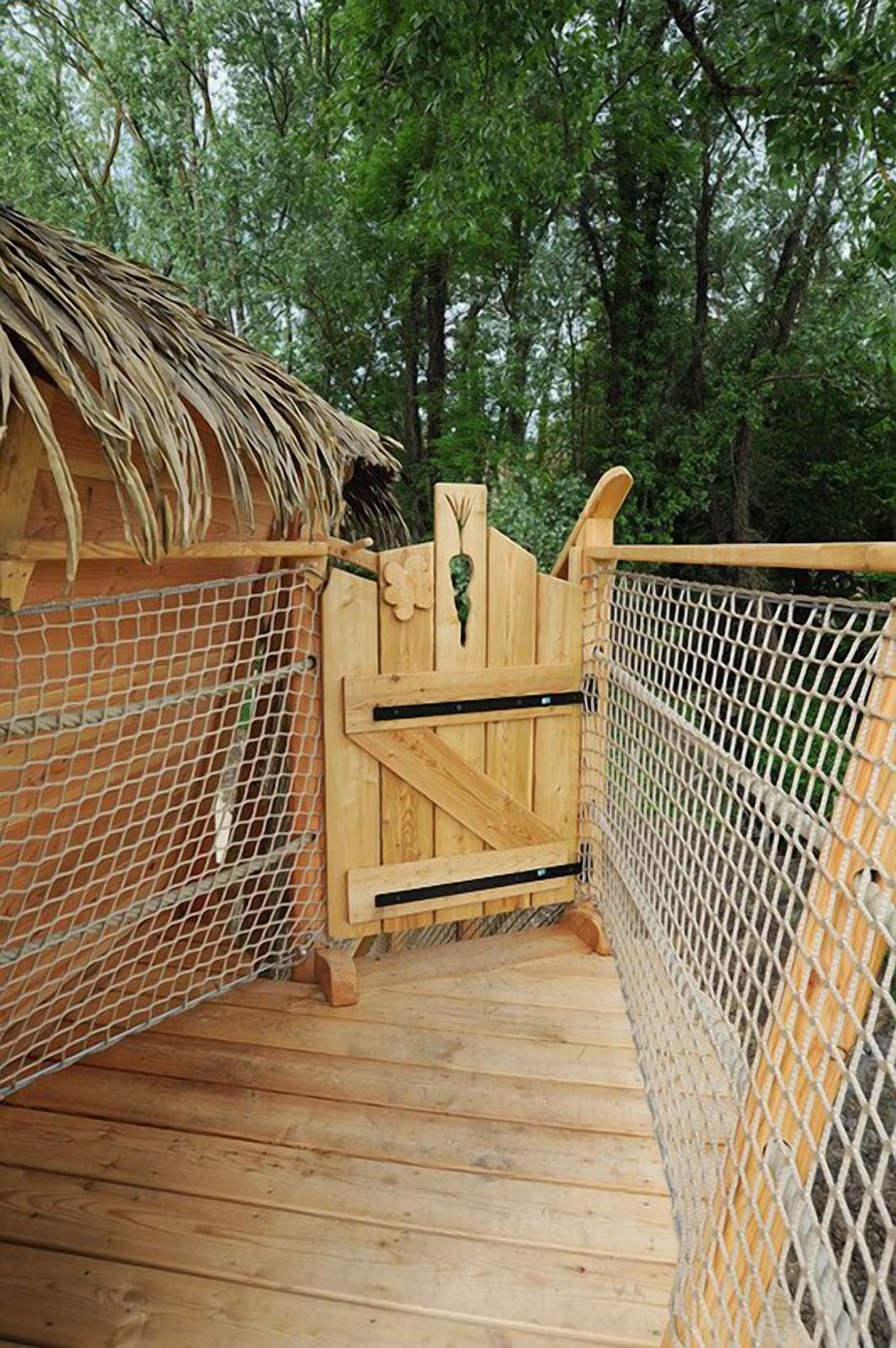 Eine Holzbrücke mit einem reetgedeckten Unterstand auf einer Seite und einem Drahtgeflechtzaun auf der anderen, die zu einem hölzernen Tor mit dekorativer Aussparung führt.