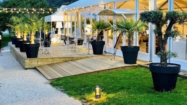 Un área de comedor exterior moderna con una terraza de madera, plantas en macetas y un edificio con cerramientos de vidrio y fachada blanca.