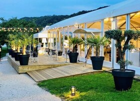 Un espace de restauration extérieur moderne, doté d’une terrasse en bois, de plantes en pot et d’un bâtiment à façade blanche, entièrement vitré.