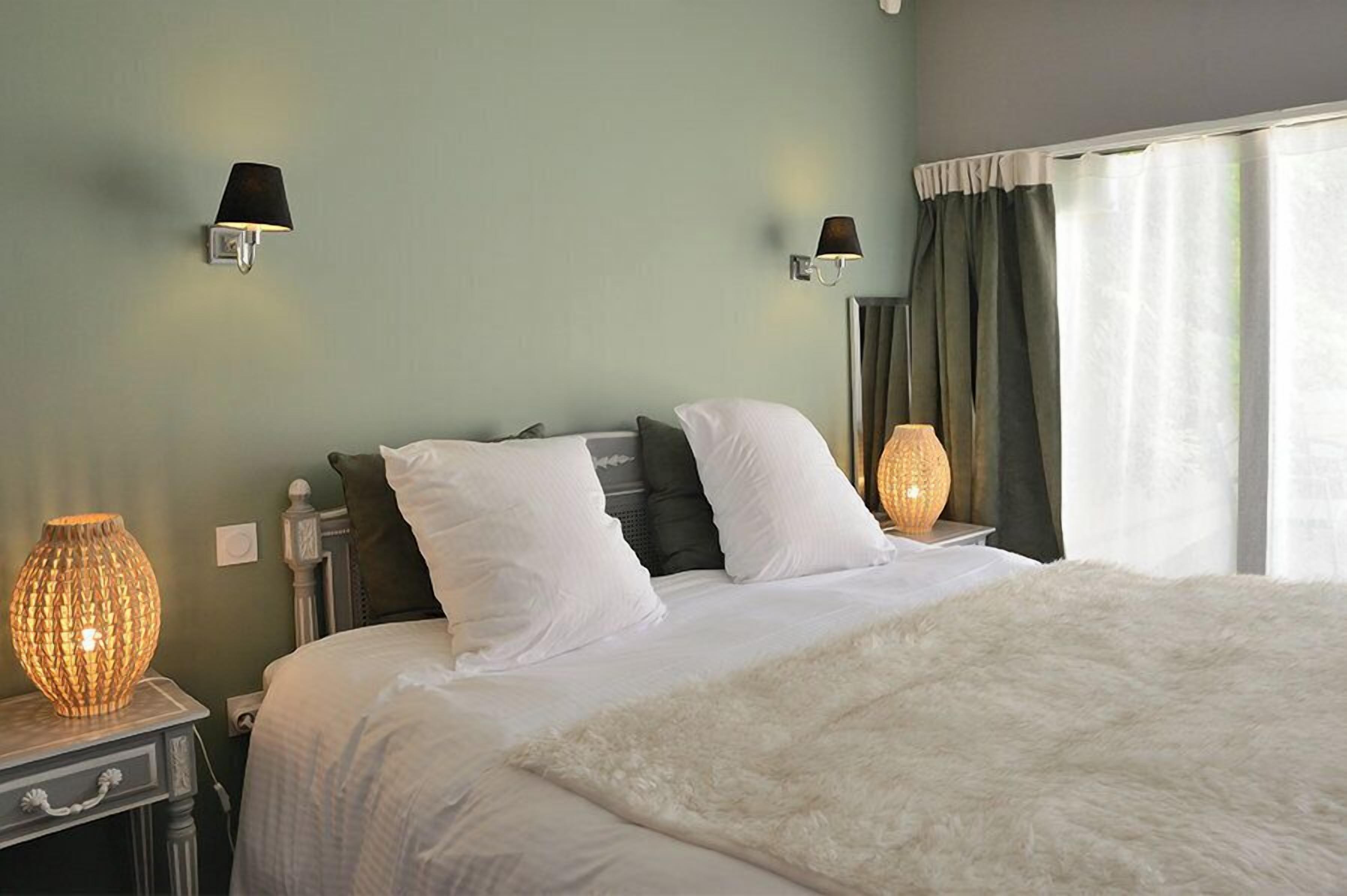 A neatly made bed with white linens and a fluffy white throw, two wall-mounted lamps, and a large window with curtains.