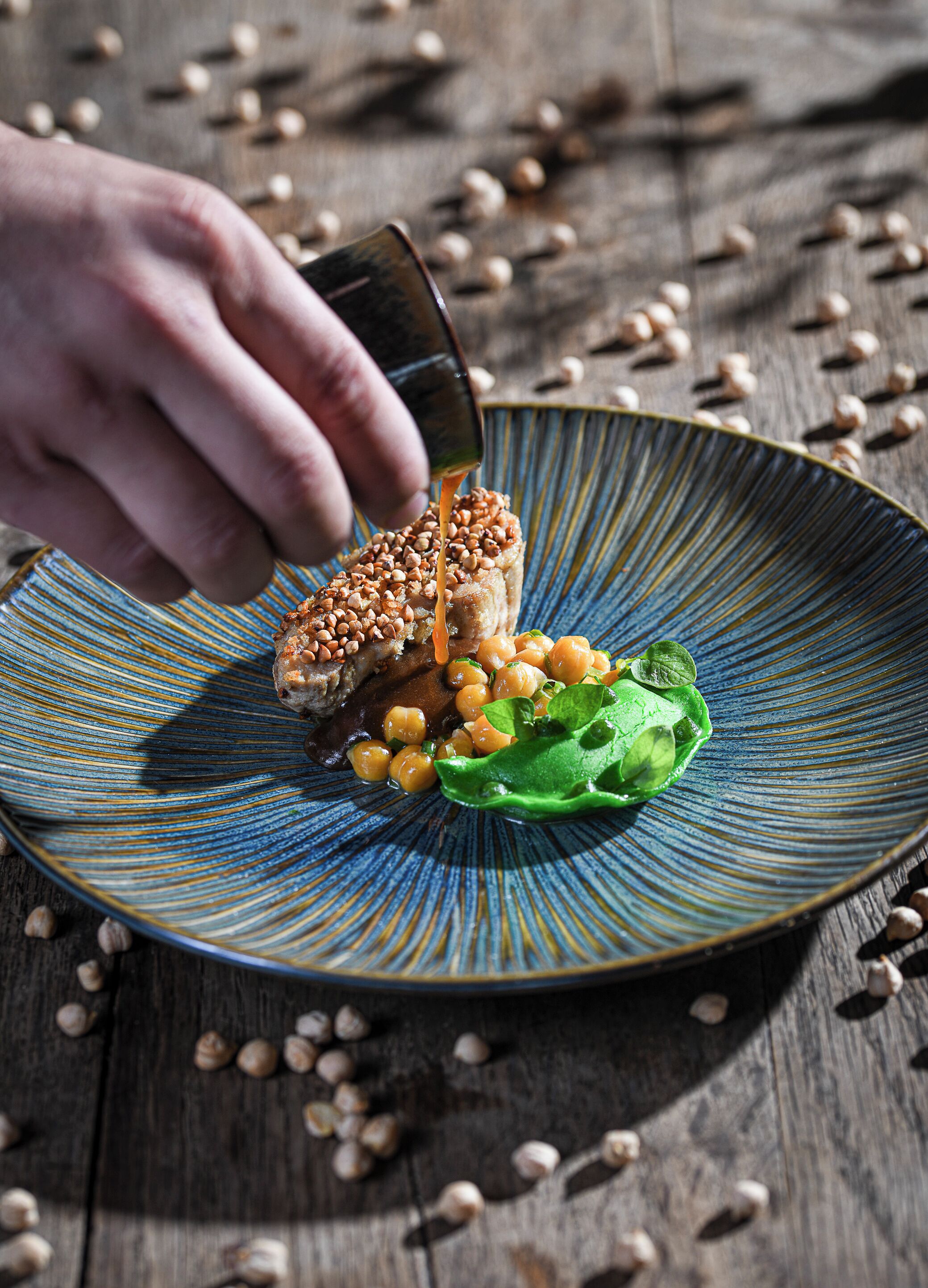 A hand pouring sauce onto a dish with chickpeas, greens, and a piece of meat on a blue-rimmed plate.