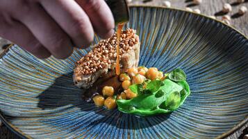 A hand pouring sauce onto a dish with chickpeas, greens, and a piece of meat on a blue-rimmed plate.