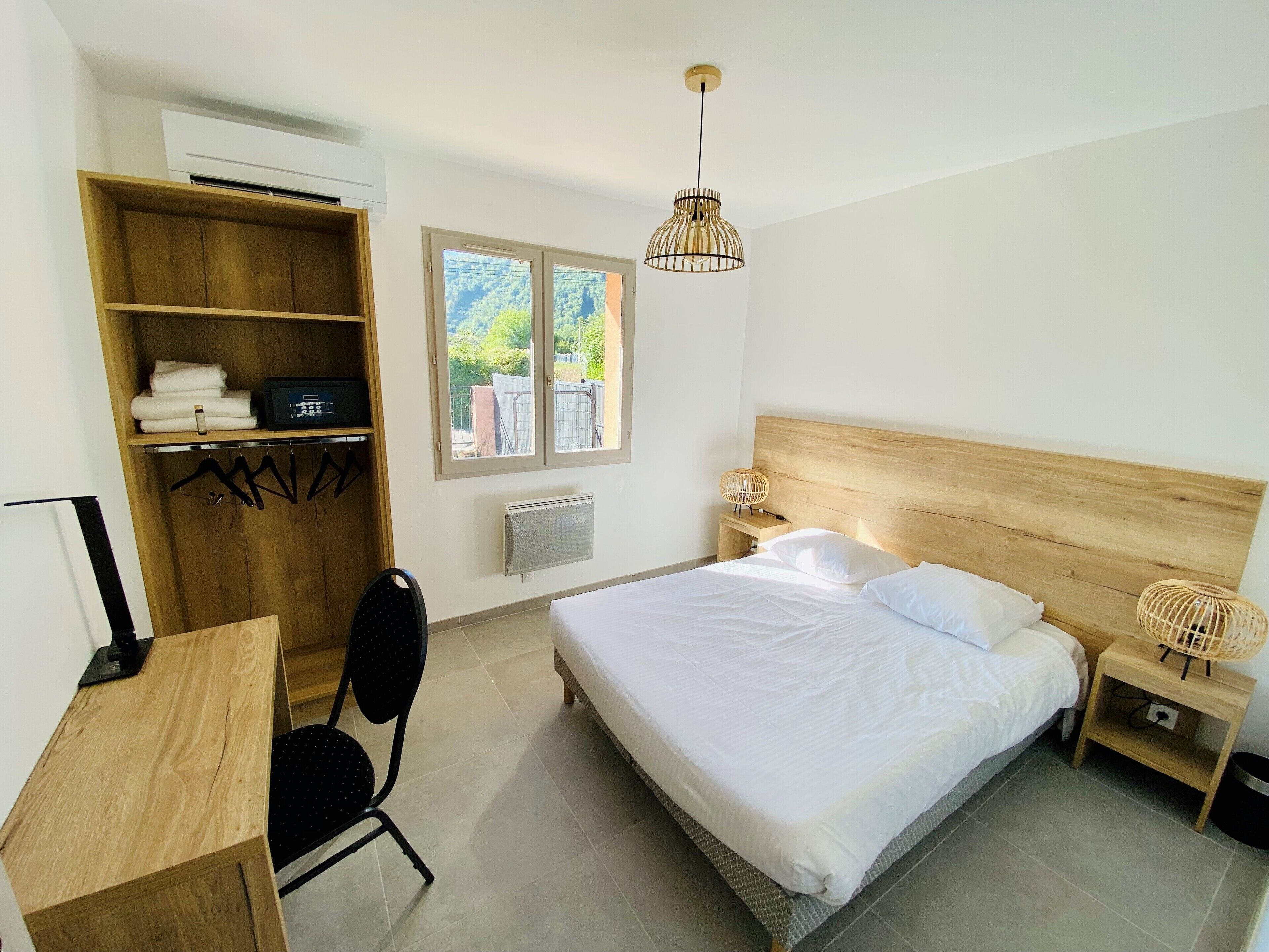 Une chambre avec un lit en bois, un bureau, une chaise, une armoire, un radiateur, une fenêtre donnant sur un paysage agréable et un luminaire suspendu.
