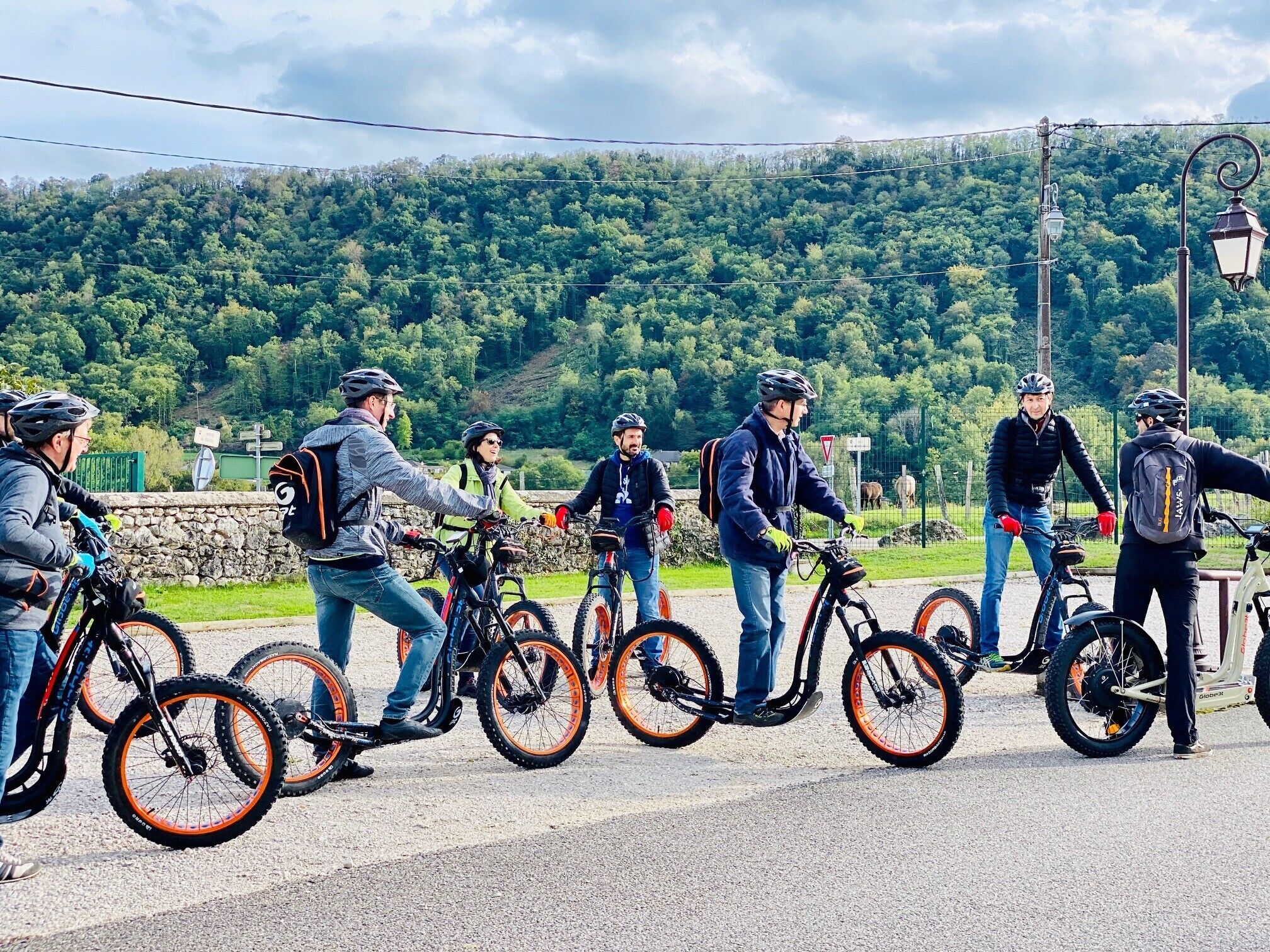 Un gruppo di persone in bicicletta su una strada, con un muro di pietra e vegetazione sullo sfondo.
