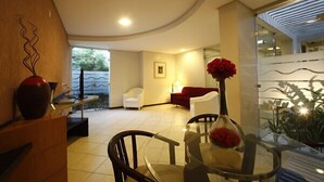 A modern living room with a glass table, chairs, a red sofa, and a vase with red flowers. - Portal da Princesa Hotel (Feira de Santana)
