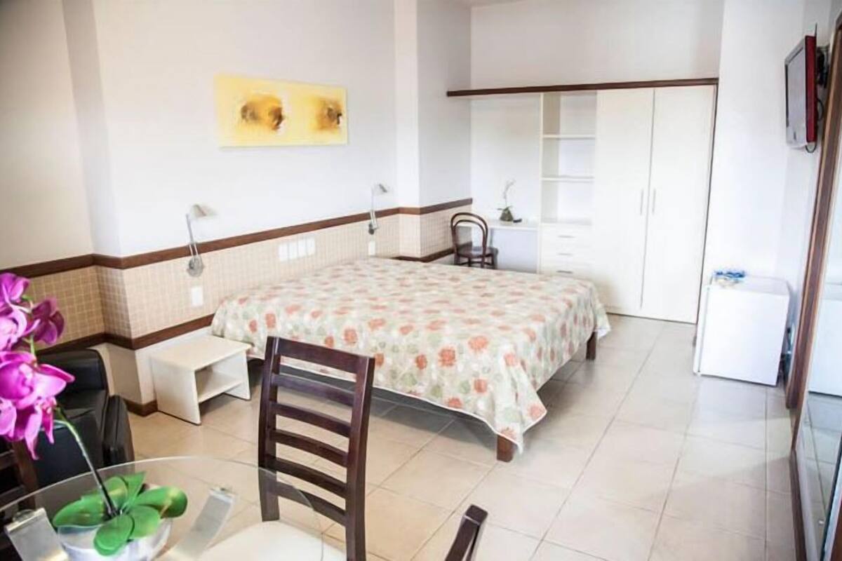 A hotel room with a bed, a desk, a chair, a small table with a plant, and a TV.