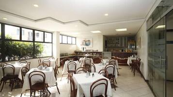 Un restaurante con varias mesas y sillas, una zona de cocina y ventanas amplias.