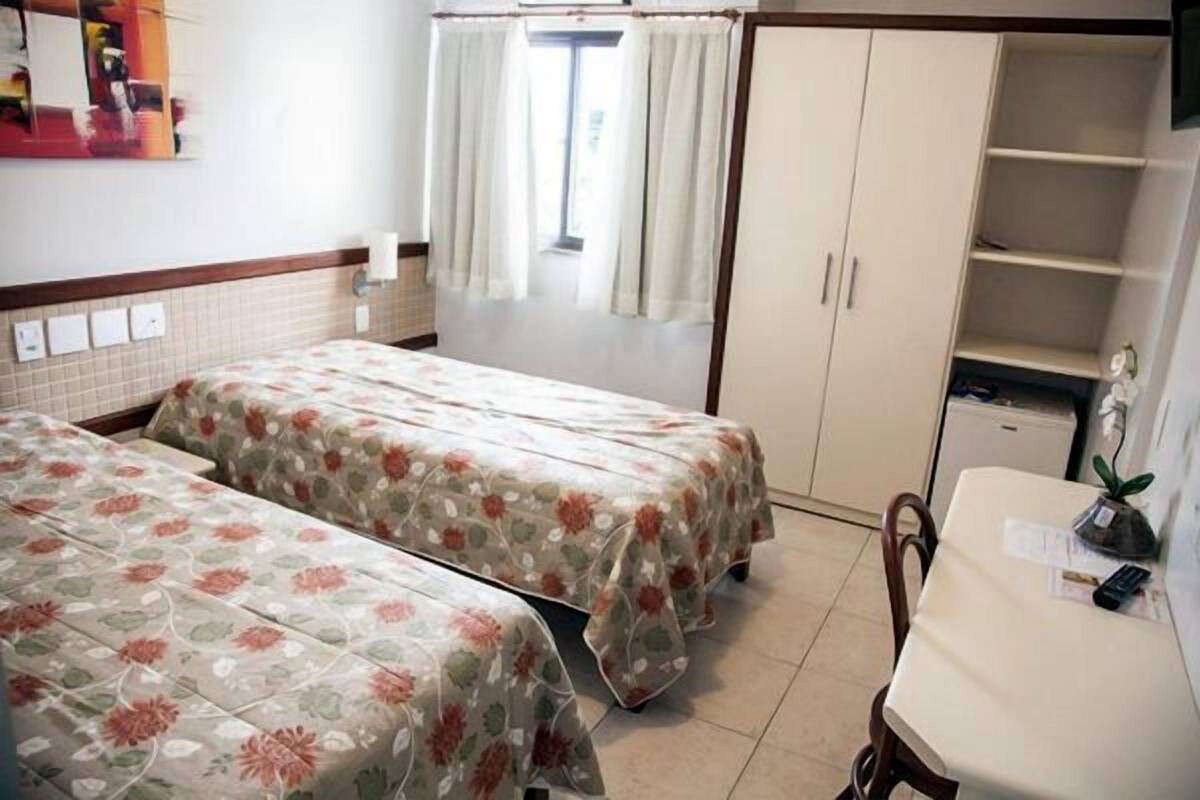 A room with two beds, a small table, a cabinet, and a window with curtains.