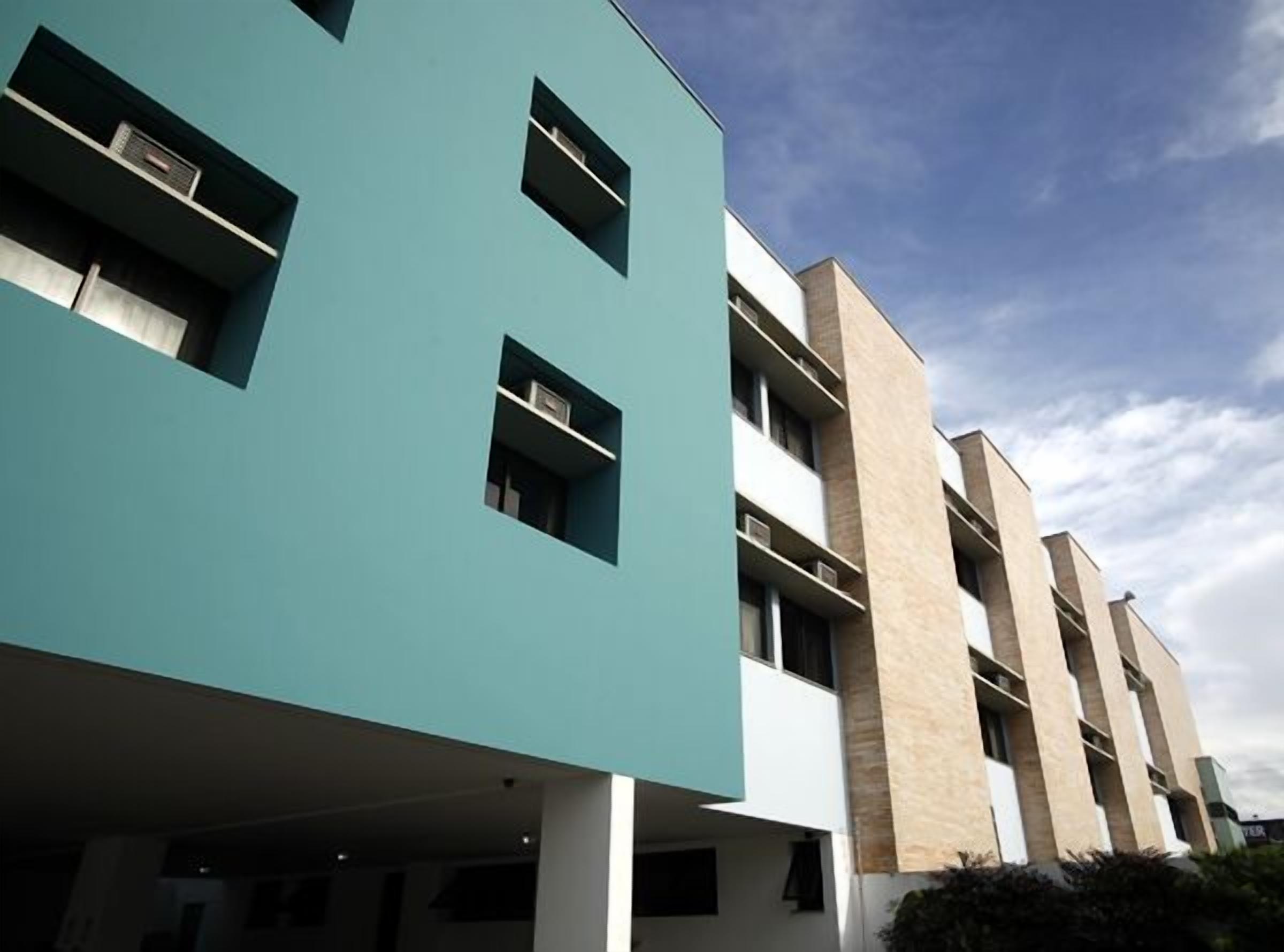 Un edificio moderno de varios pisos con fachada plana, grandes ventanales y una combinación de tonos beige y turquesa.