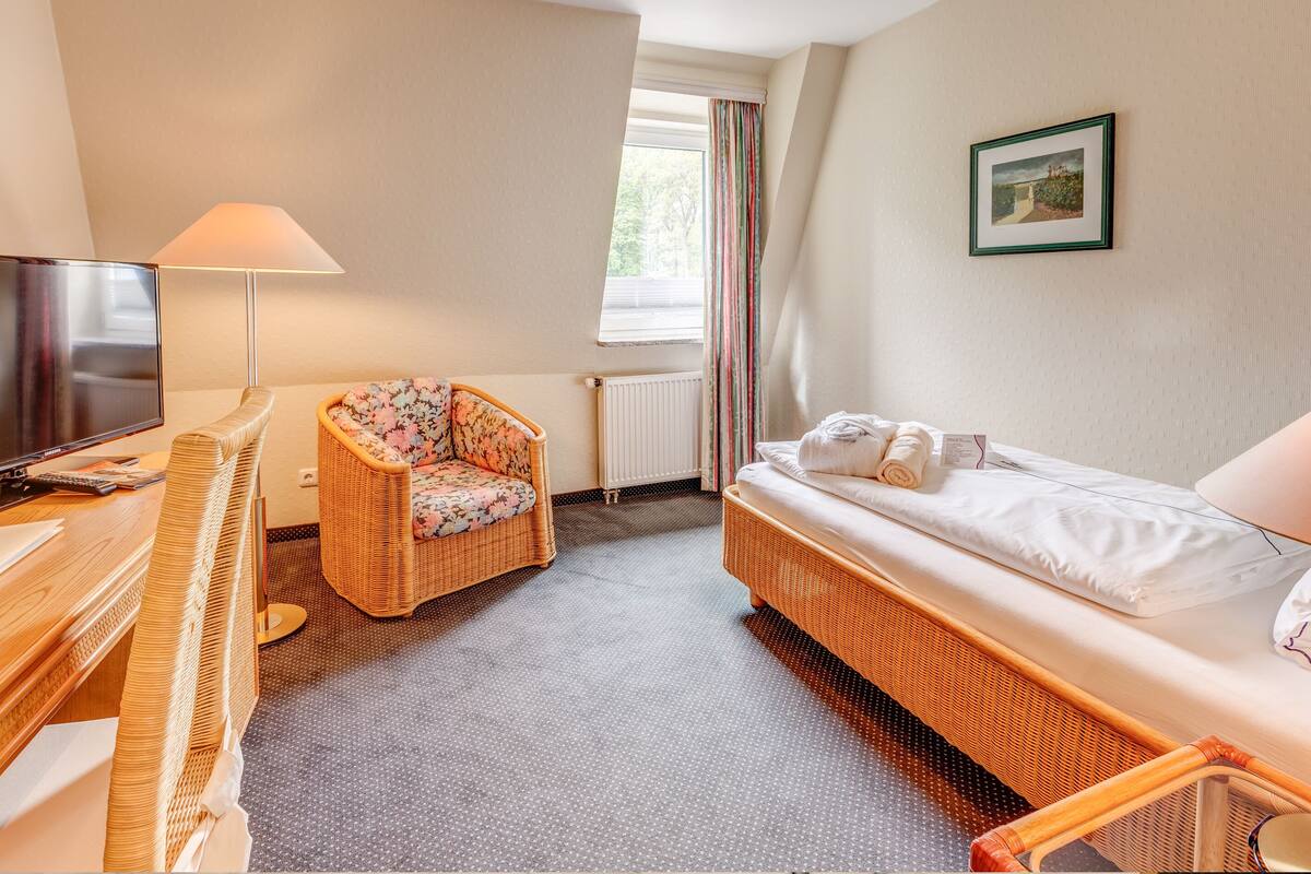 A cozy room with a bed, a wicker chair, a desk with a lamp, and a television.