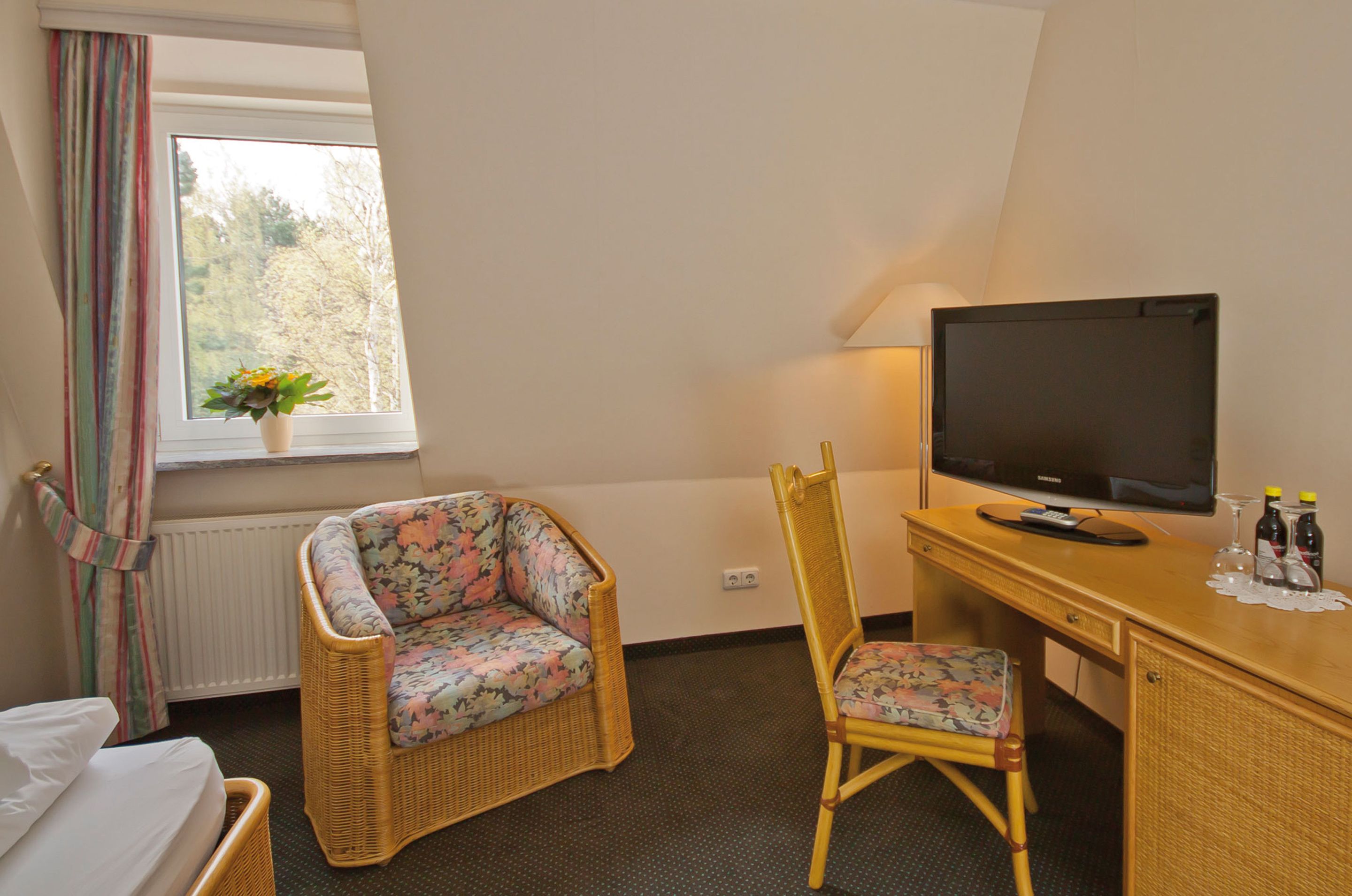 A hotel room with a bed, a wicker chair, a desk with a television, and a window with a view of trees.