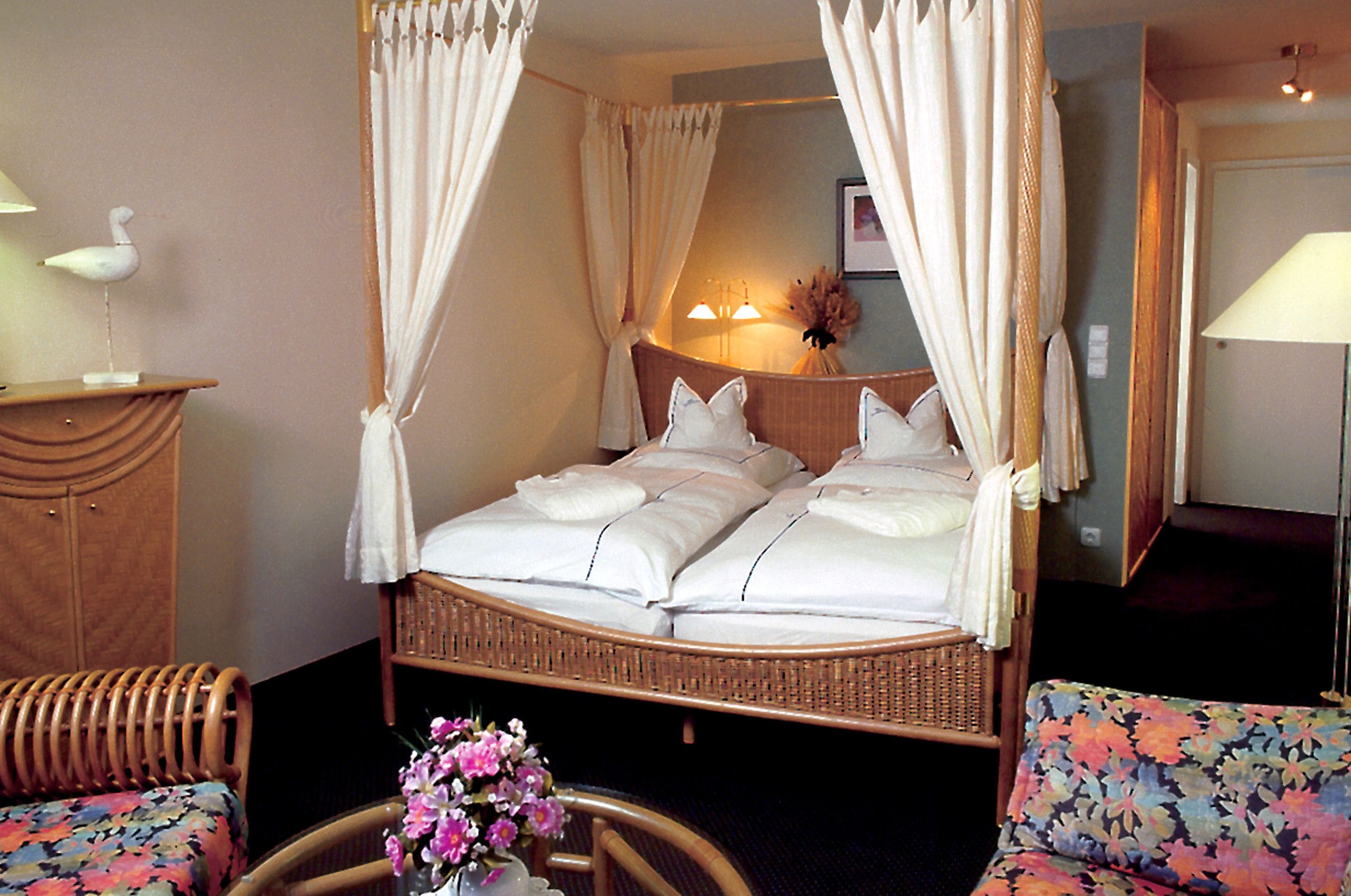 Una cama con dosel, una lámpara de noche, un sillón floral y una mesita con flores.