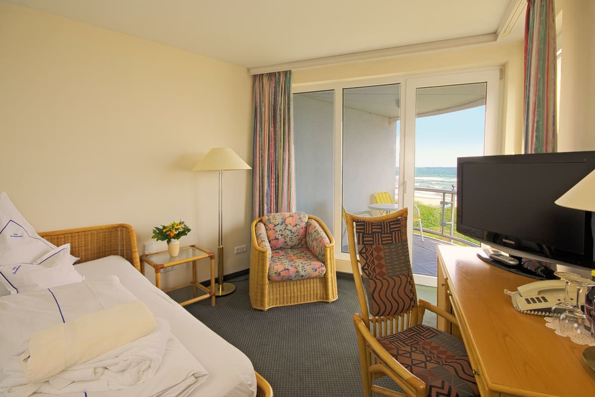 A hotel room with a bed, a television, a chair, a small table with a lamp, and a view of the ocean through a sliding door.
