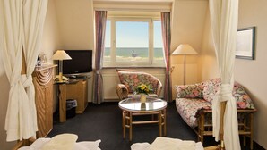 Habitación de hotel con cama, sofá, sillón, mesa, televisión y ventana con vista al mar.