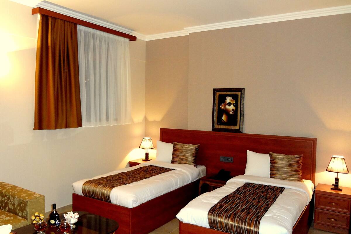 Ein Hotelzimmer mit zwei Betten, einem Sofa, einem Tisch mit Flasche und Gläsern sowie einem Gemälde an der Wand.