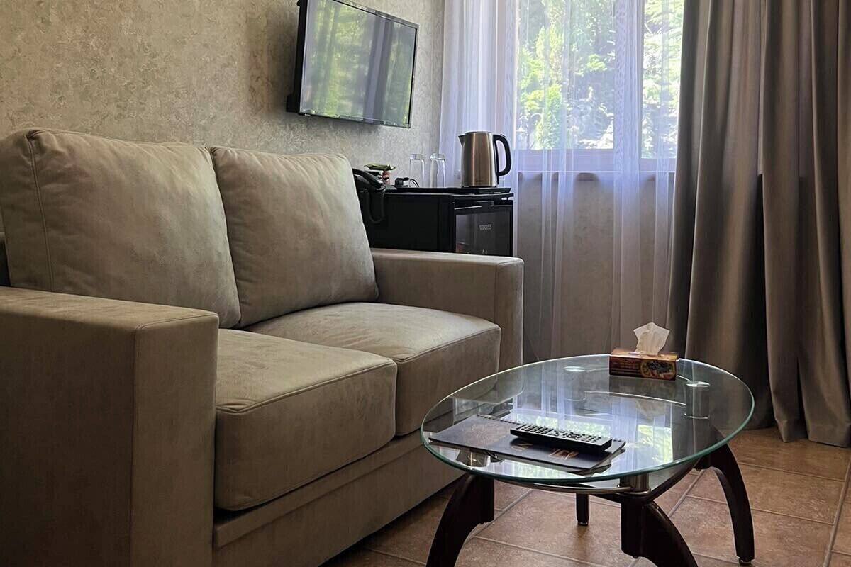 Ein Wohnzimmer mit einem beigen Sofa, einem Glastisch, einem an der Wand montierten Flachbildfernseher und einem Fenster mit Vorhängen.