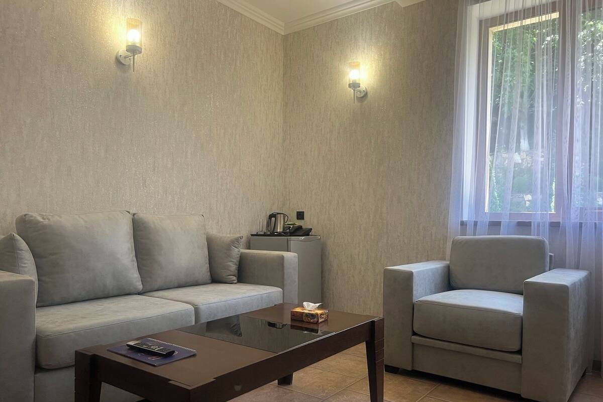 Ein Wohnzimmer mit einer Couch, einem Sessel und einem Couchtisch. Zwei Wandleuchten und ein Fenster mit durchsichtigen Vorhängen sind ebenfalls vorhanden.