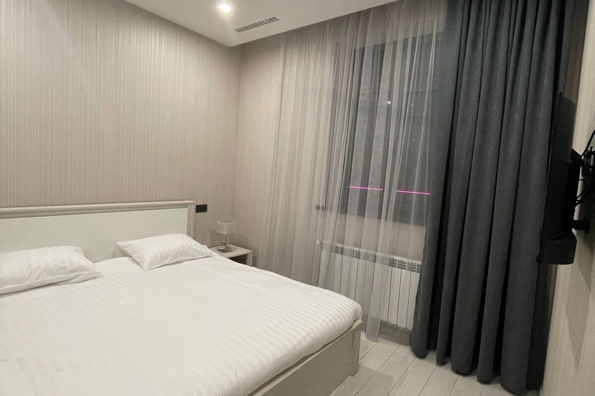 Ein modernes Schlafzimmer mit einem großen Bett, einem Nachttisch, einem an der Wand montierten Fernseher und einem Fenster mit leichtem Vorhang.