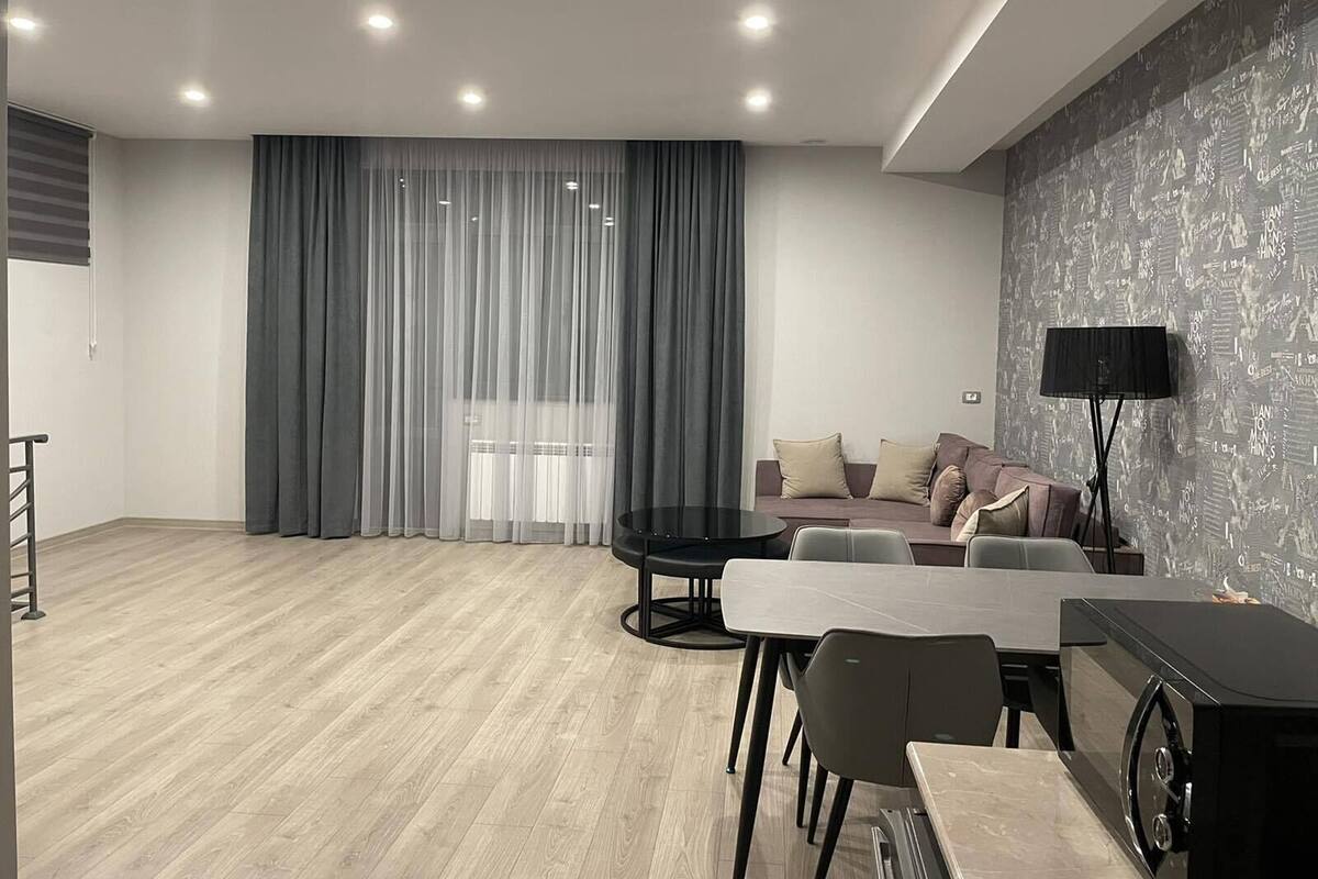 Ein modernes Wohnzimmer mit Essbereich, grauen Vorhängen, einer Couch mit Kissen, einem Flachbildfernseher und einer schwarzen Stehlampe.