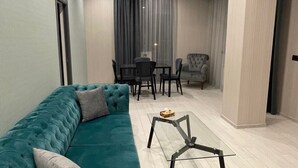 Una sala moderna con un sofá color turquesa, una mesa de centro y un reposapiés gris. Se observa un comedor con mesa y sillas al fondo.