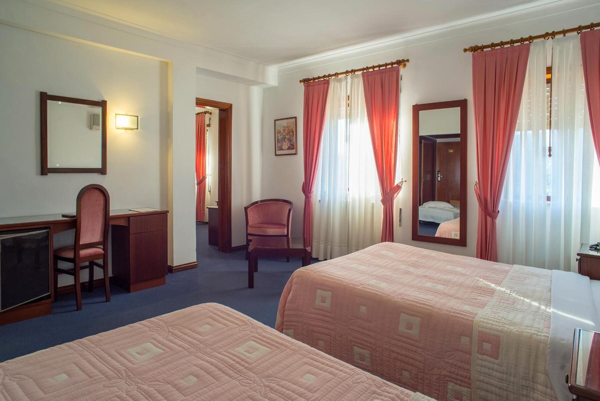 A hotel room with two beds, a desk, a chair, and a mirror.