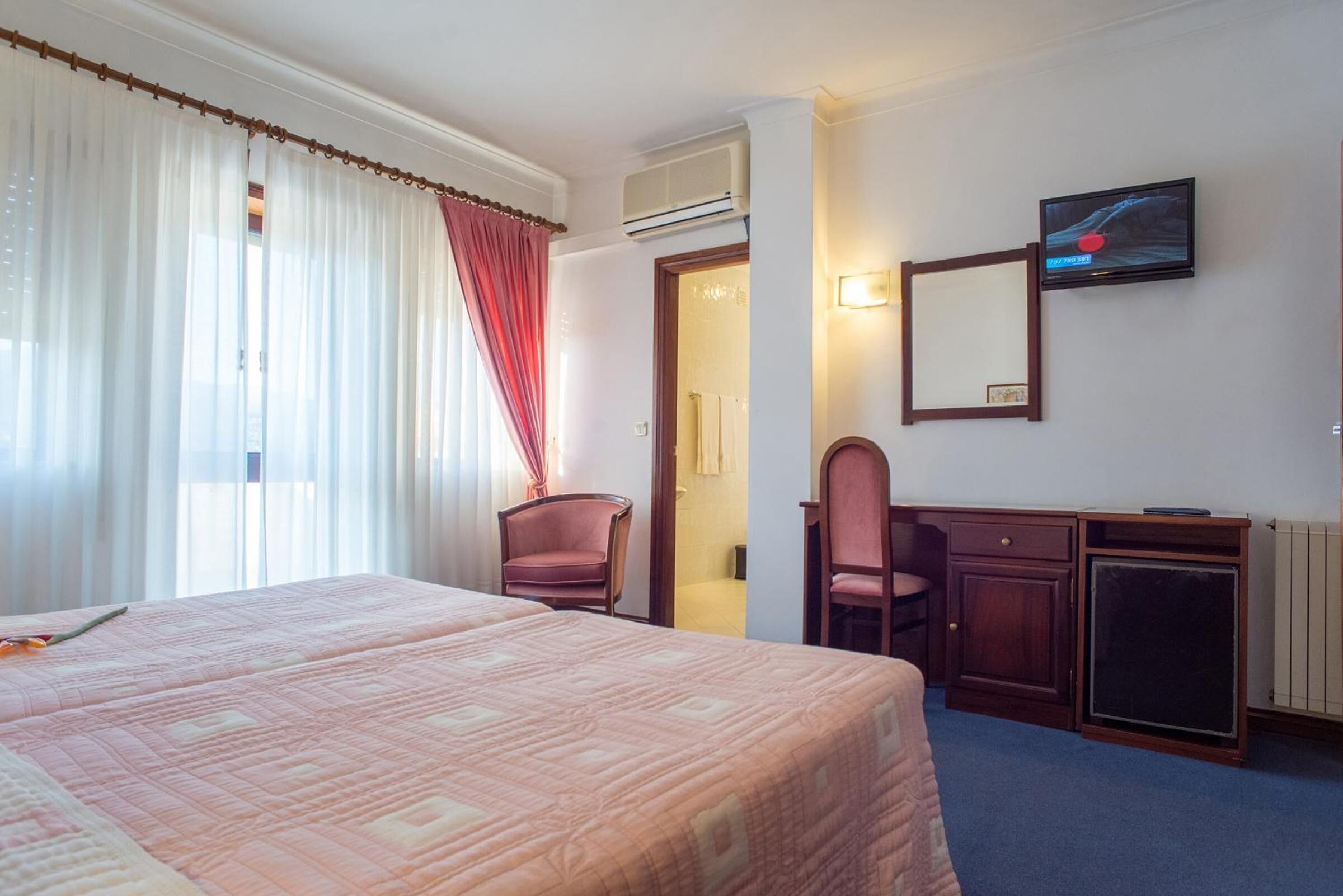 Habitación de hotel con una cama grande, un escritorio con una silla, televisión y ventana con cortinas.