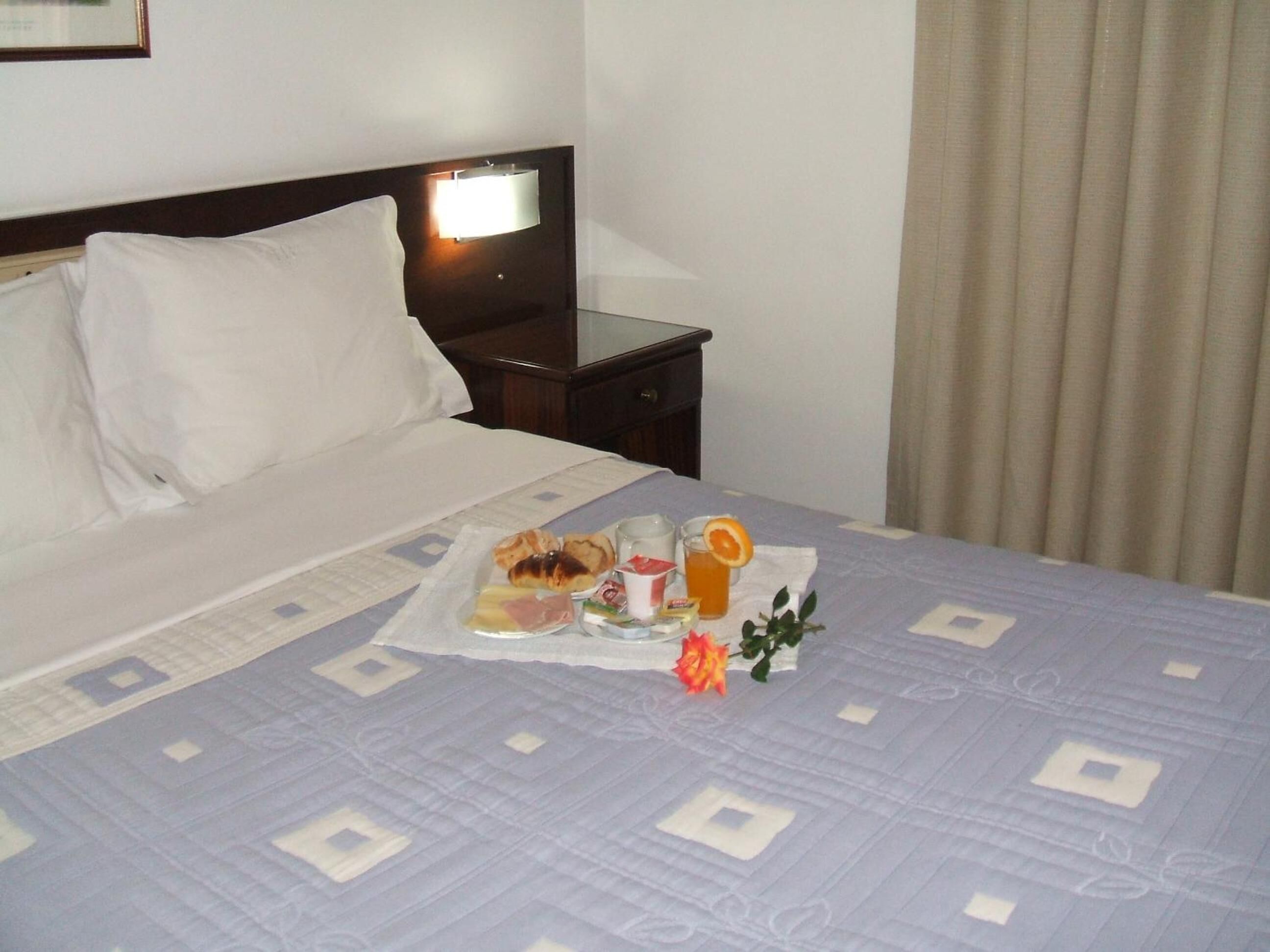 Habitación de hotel con una cama bien hecha, una mesita de noche con lámpara y una bandeja de desayuno con cruasanes, fruta y una bebida.