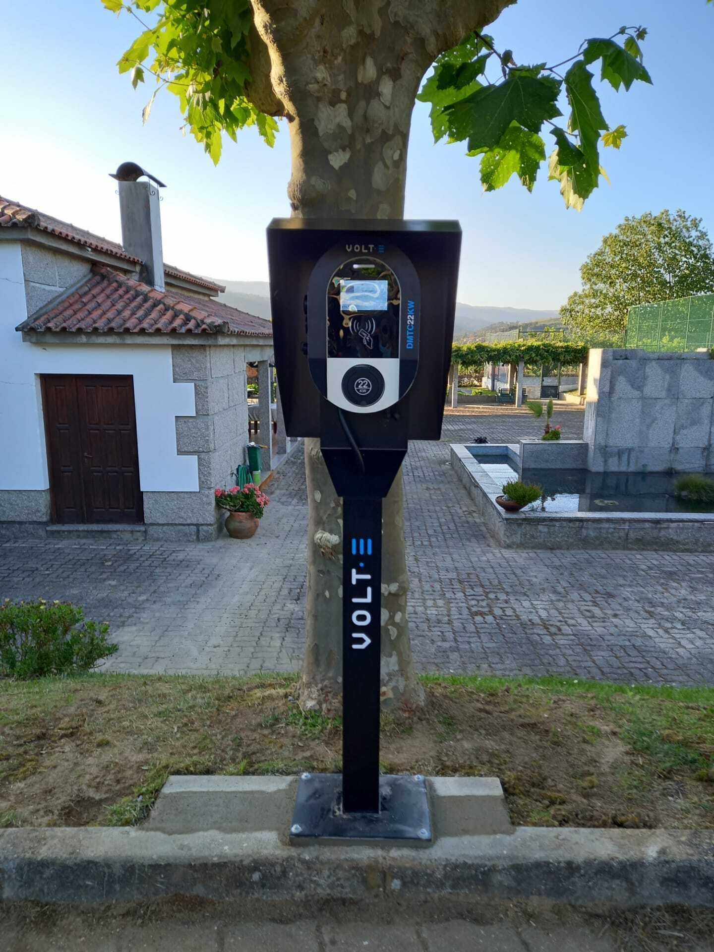 A public electric charging station with a digital display, located in a courtyard with a tree, a building with a tiled roof, and a stone wall.