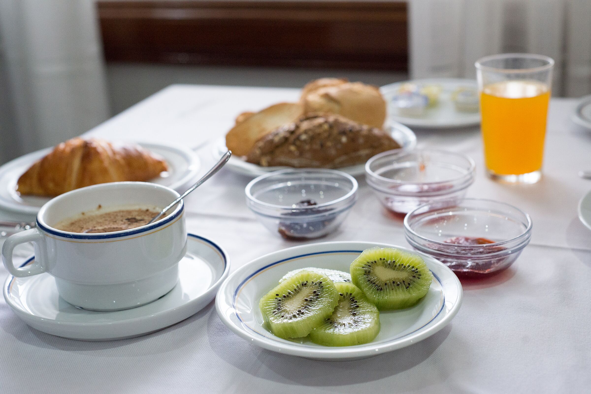 A breakfast setting with a croissant, kiwi slices, a cup of coffee, and a glass of orange juice on a table.