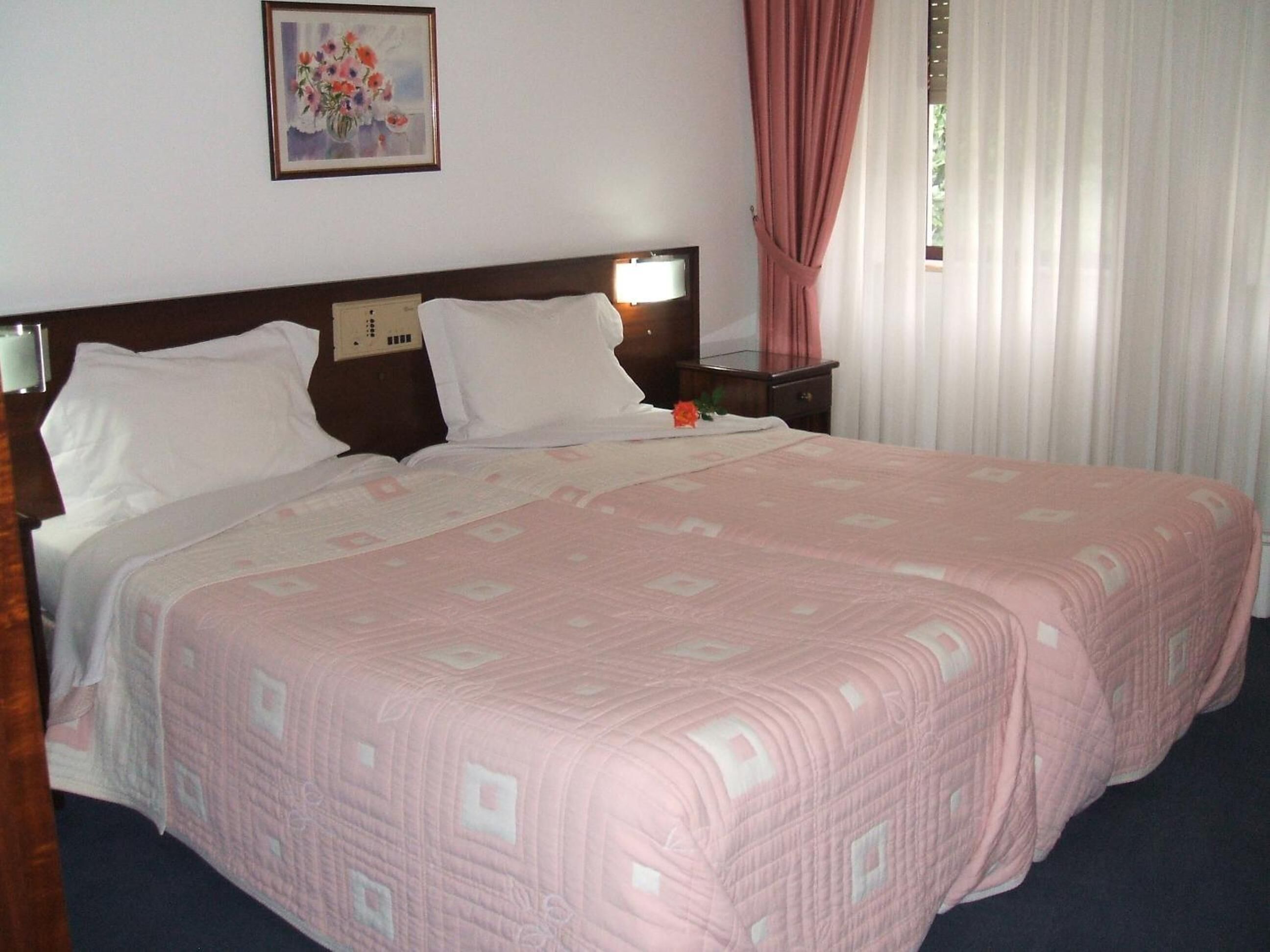 Cama doble con colcha rosa, mesita de noche, cuadro floral enmarco y ventana con cortinas traslúcidas.