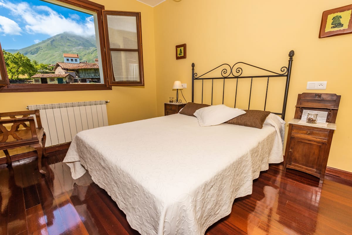 A bedroom with a wooden floor, a bed with white linens, a nightstand with a lamp, a window with a view of houses and a mountain, and a small wooden cabinet.
