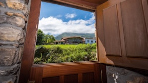 A rustic stone wall and wooden door leading to a view of a traditional building with a thatched roof, surrounded by lush greenery and mountains in the background.