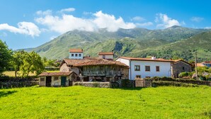 A rural village with traditional stone and tiled-roof buildings, surrounded by lush green fields and mountains in the background.