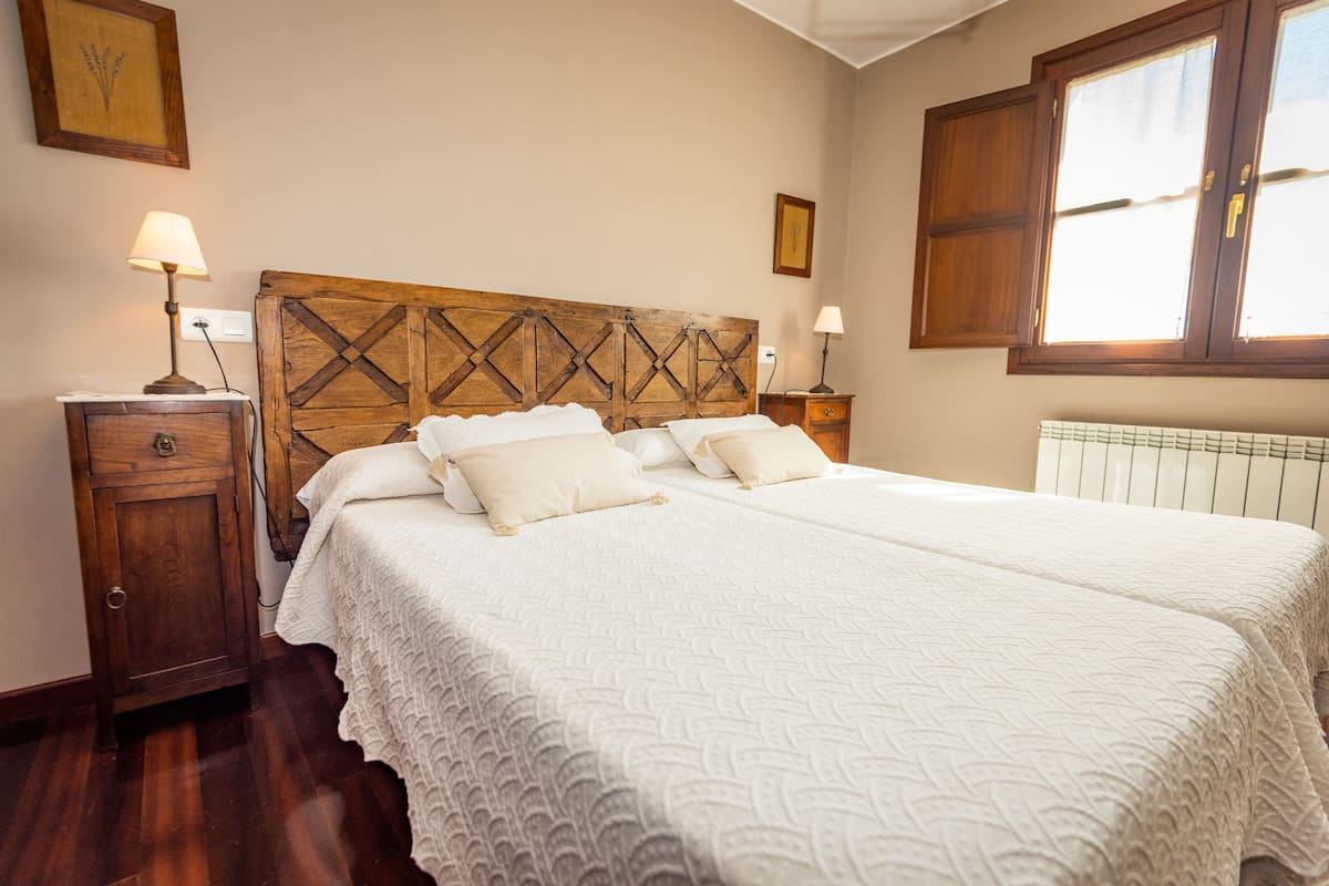 A bedroom with a wooden headboard, two bedside tables with lamps, a large bed with white bedding, and a window with wooden shutters.