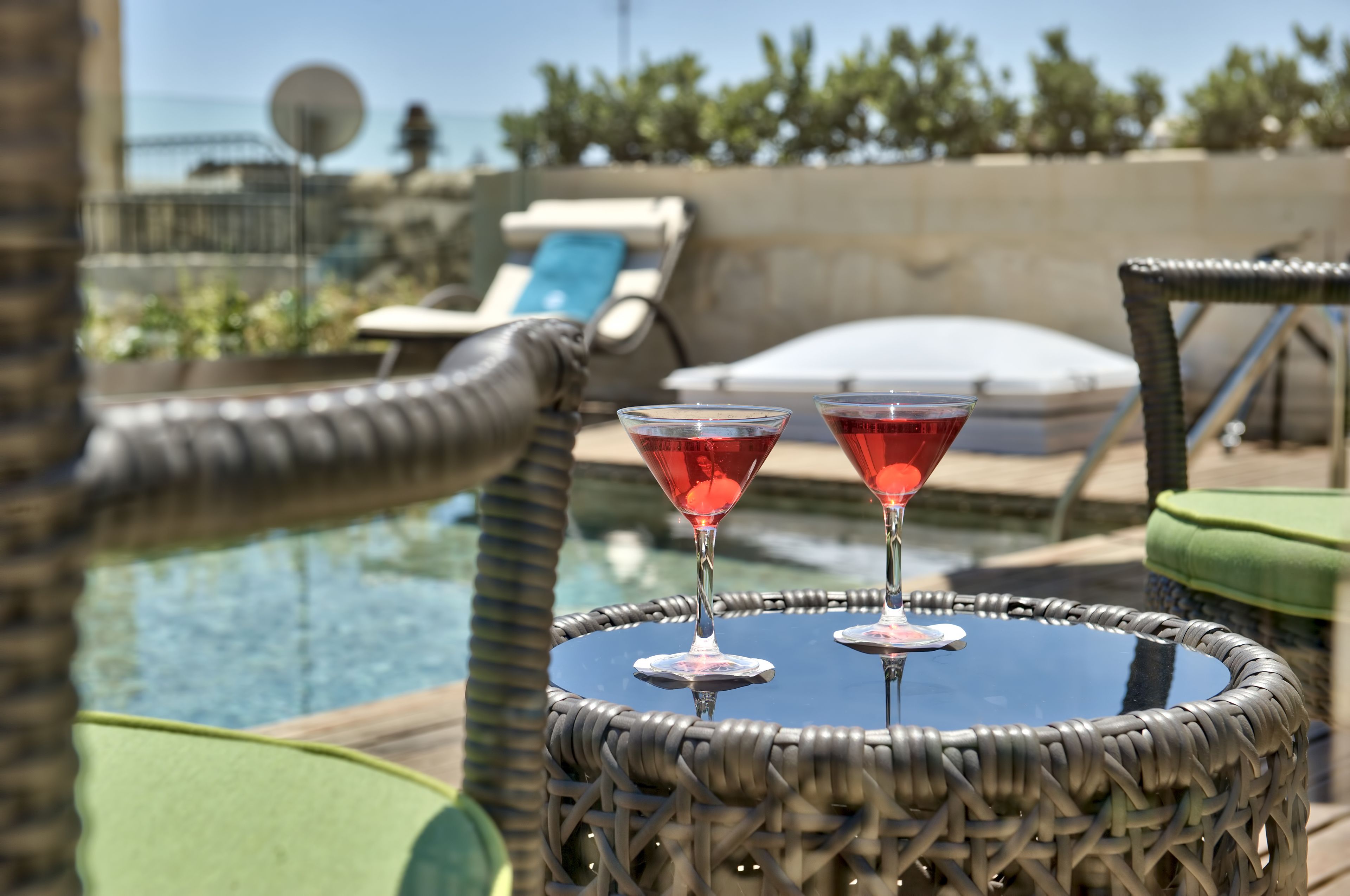 A wicker table with two martini glasses filled with red liquid, positioned near a poolside area with lounge chairs and a view of the sea.