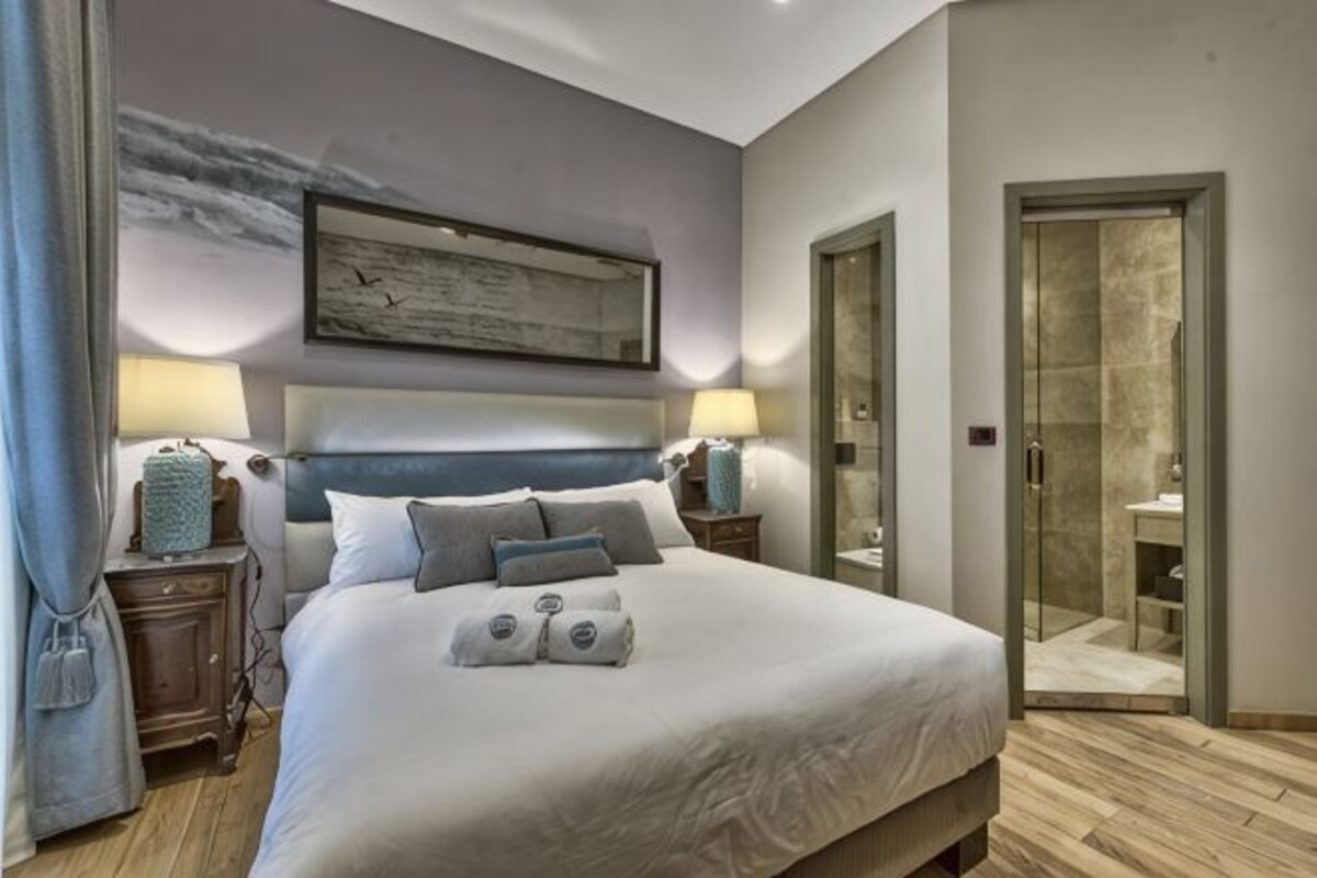 A bedroom with a large bed, bedside lamps, a painting on the wall, and a bathroom visible through an open door.