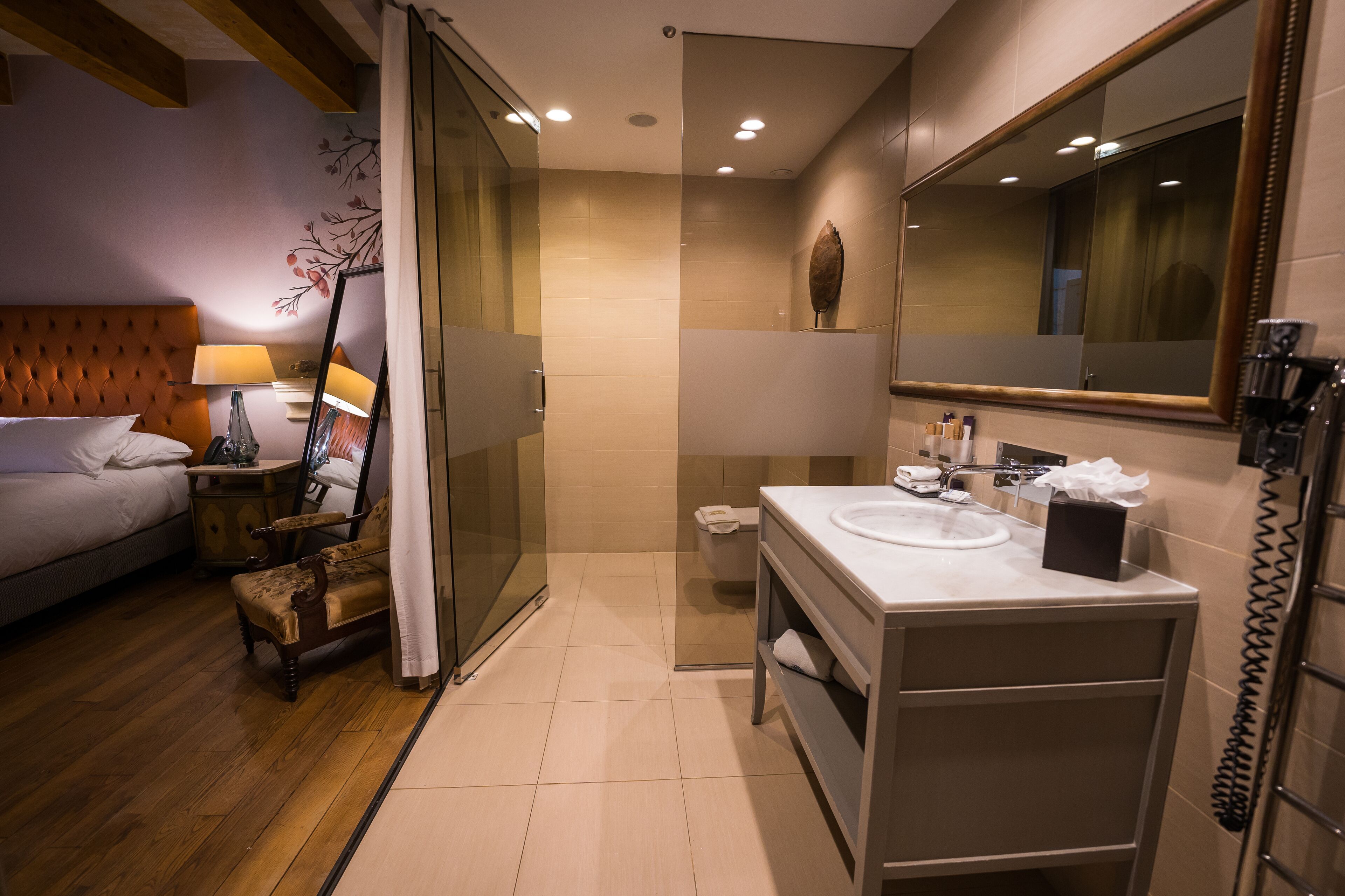 Un baño de hotel moderno con cabina de ducha de vidrio, una silla de madera, una mesita de noche con lámpara y un espejo grande.