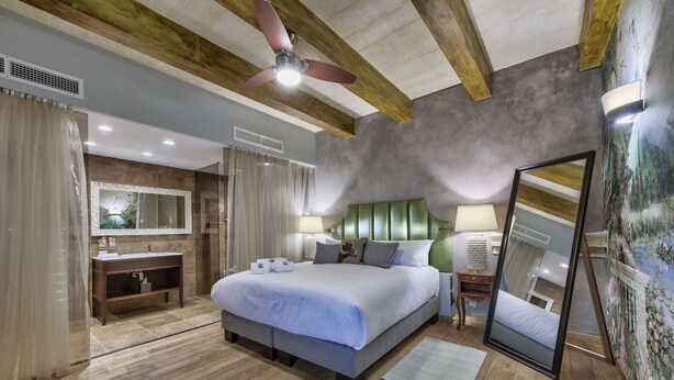 A modern bedroom with a large bed, wooden beams, a ceiling fan, a vanity with a mirror, and a nightstand with a lamp.