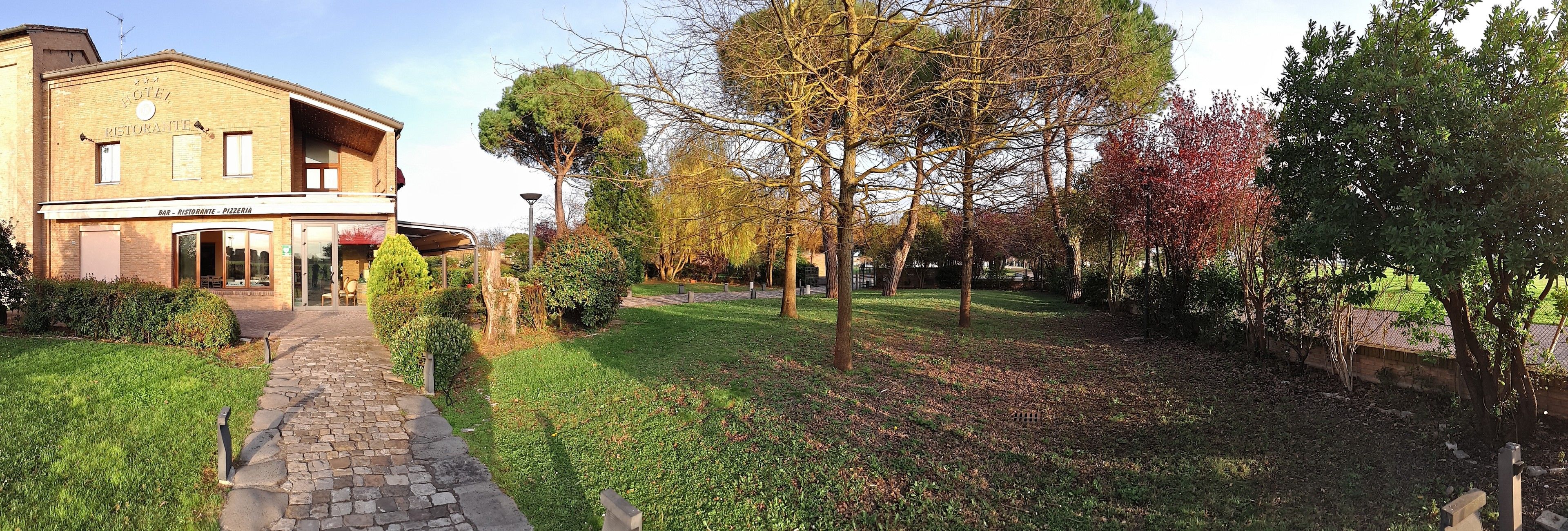 A hotel with a bar and restaurant on the ground floor, a stone pathway leading to the entrance, and a well-maintained garden with trees and shrubs.