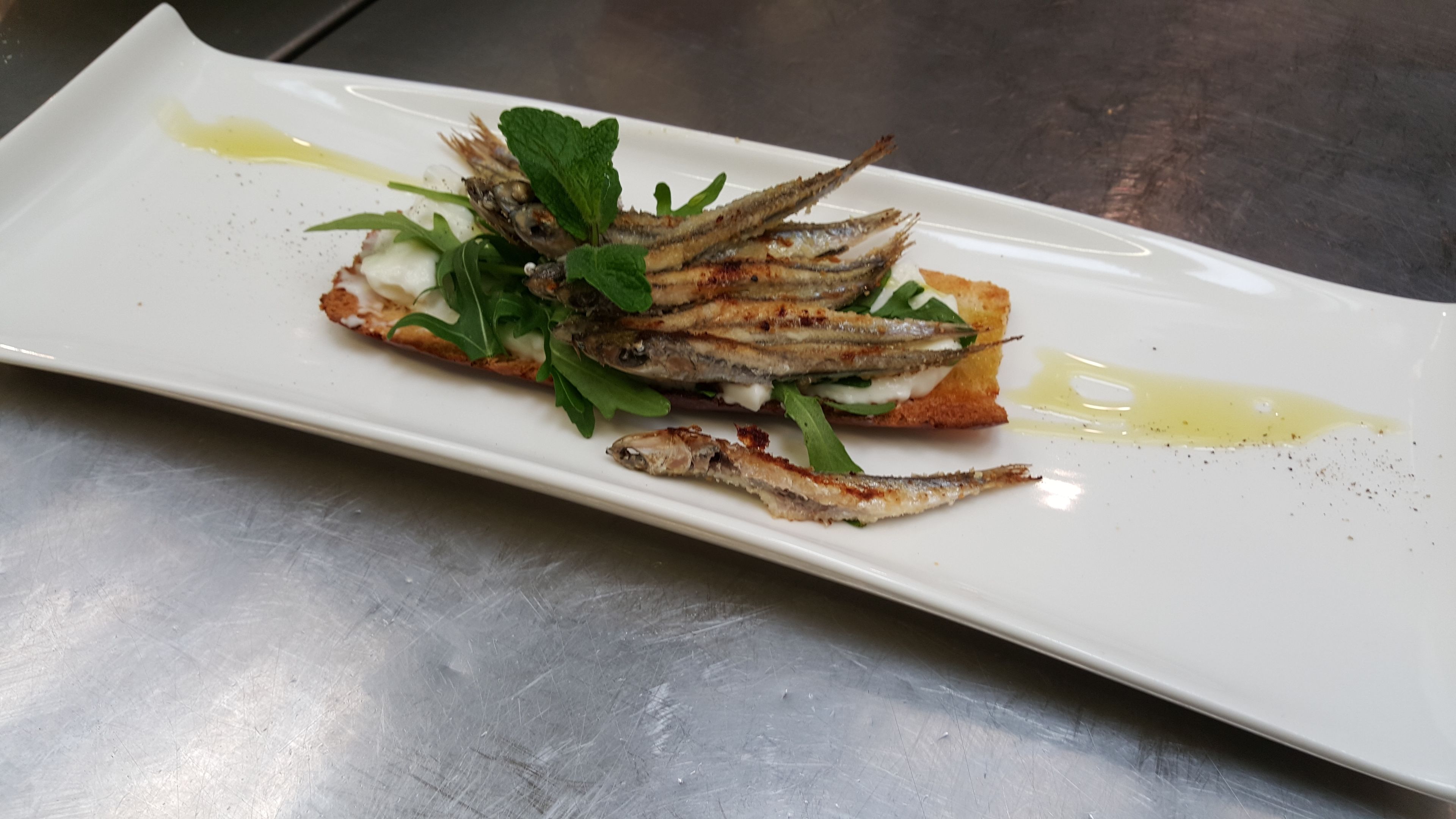 Un piatto rettangolare bianco con pesce alla griglia, verdure e salsa cremosa, su una superficie metallica.