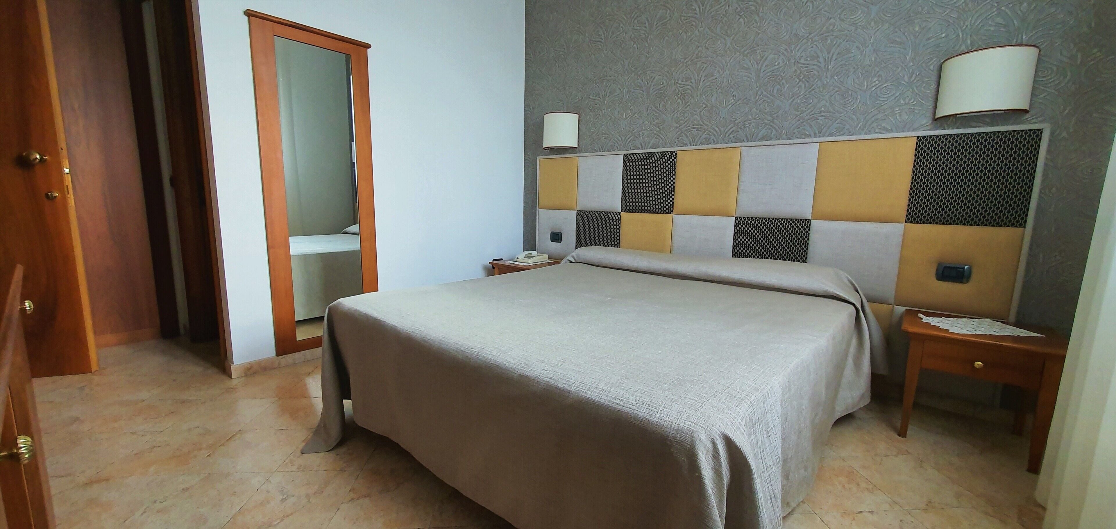Quarto de hotel com cama, mesa de cabeceira, abajur e guarda-roupa.