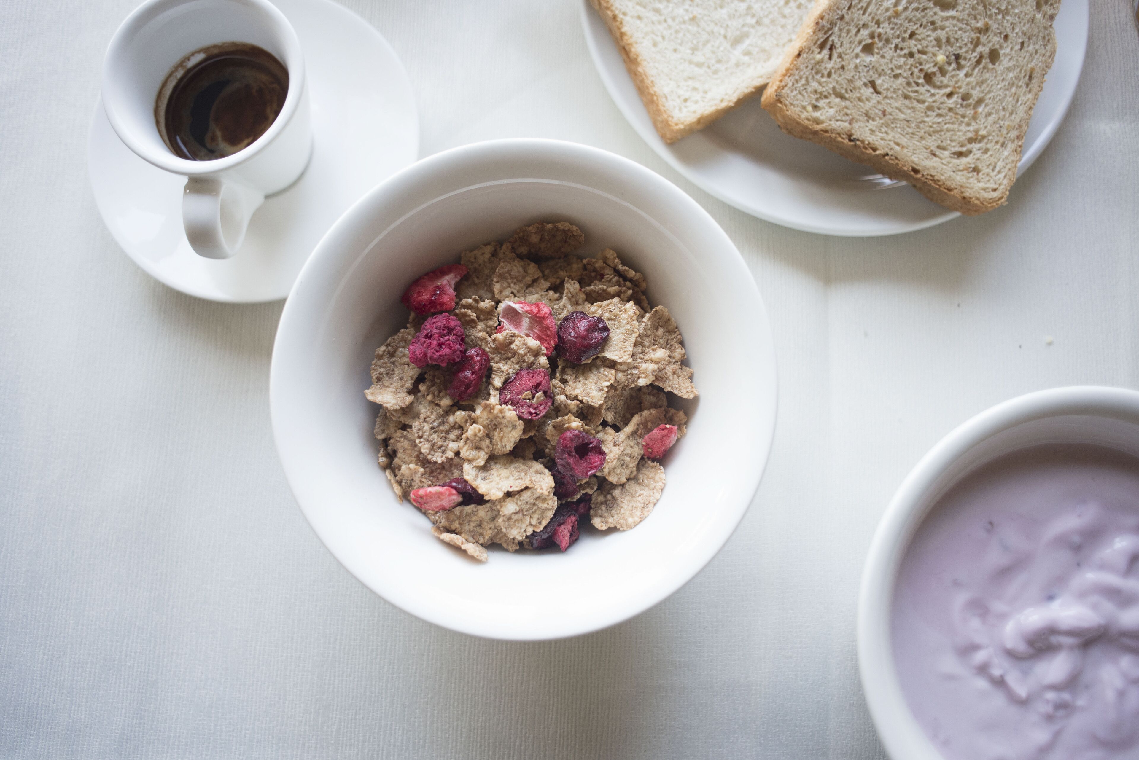 A bowl of cereal with berries, a cup of coffee, and slices of bread on a white table.