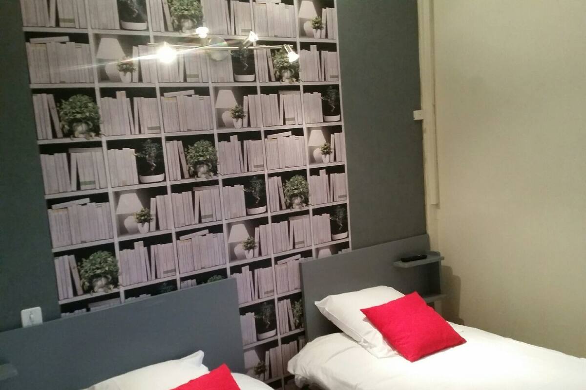 A room with two beds, each with white bedding and a red pillow. A large bookshelf filled with books and plants is mounted on the wall.