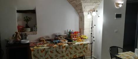 Uma área de refeições com mesa de buffet coberta por uma toalha florida, parede de pedra e teto com tijolos aparentes.