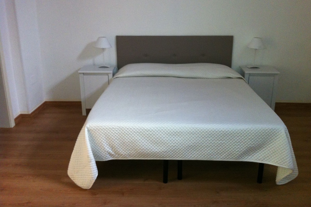 Una cama bien hecha con colcha blanca en una habitación sencilla con piso de madera y paredes blancas.