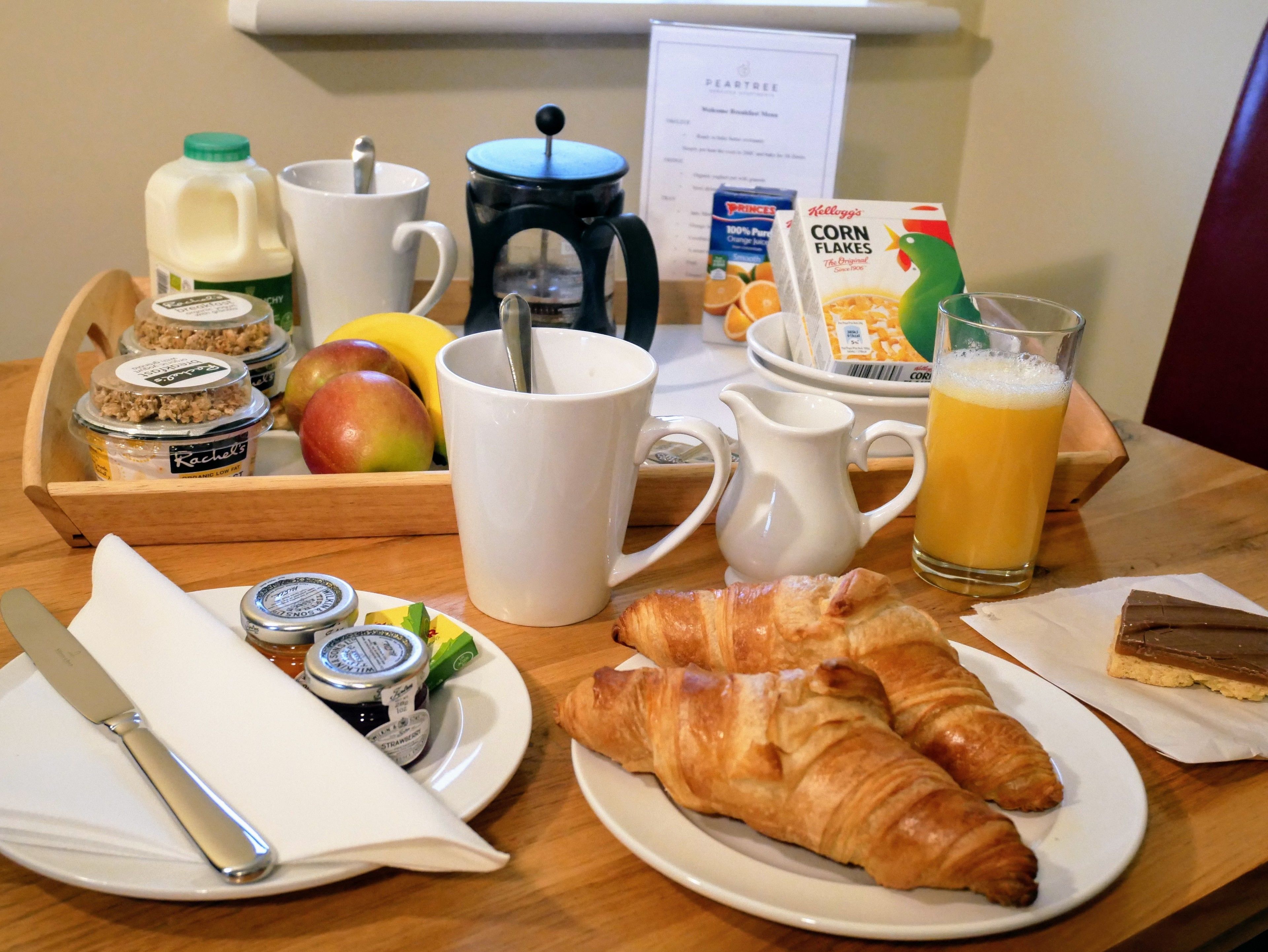 A breakfast tray with croissants, a glass of orange juice, a coffee pot, a milk jug, a fruit bowl, and cereal boxes.