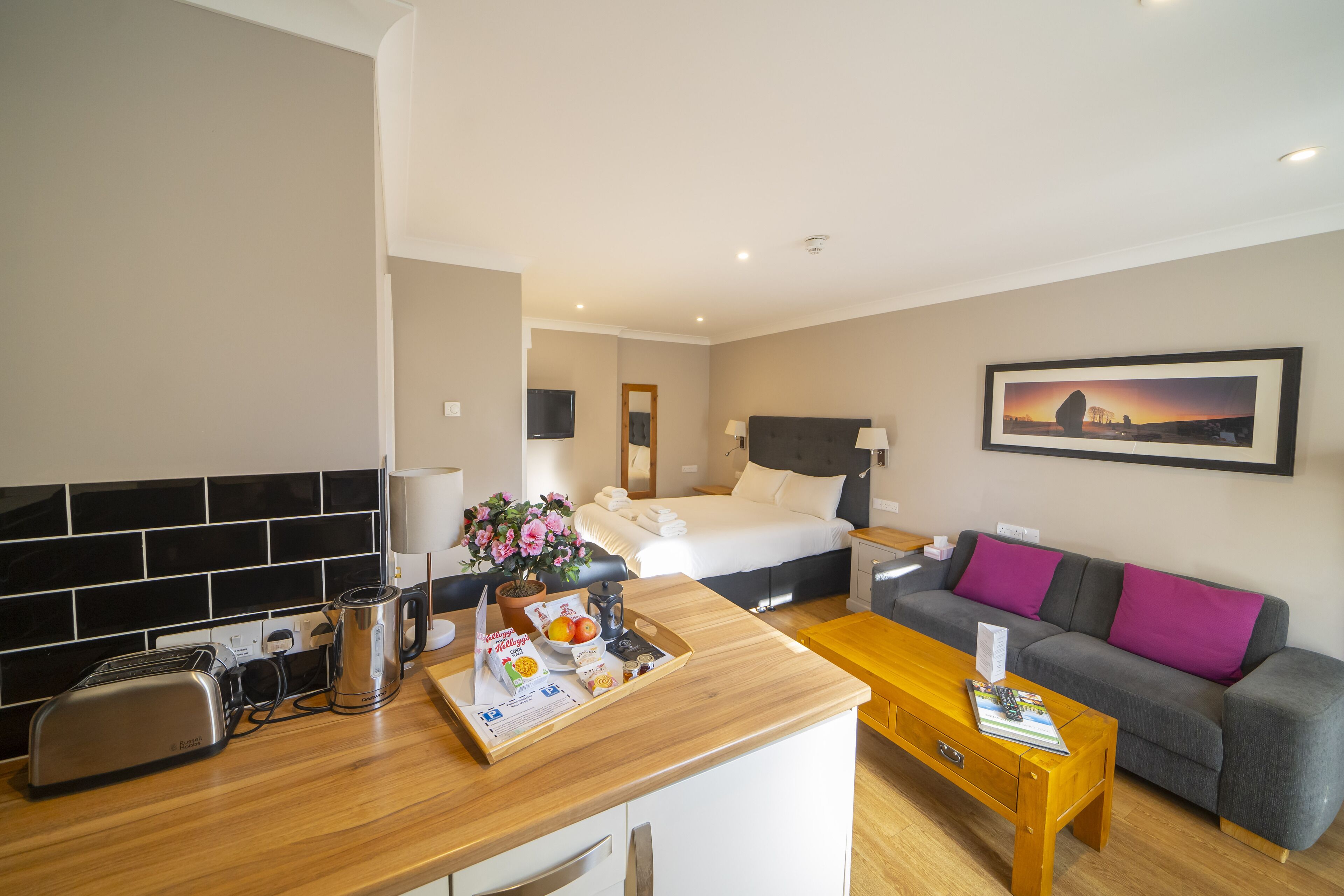 A hotel room with a kitchenette, a bed, a sofa, and a painting on the wall.