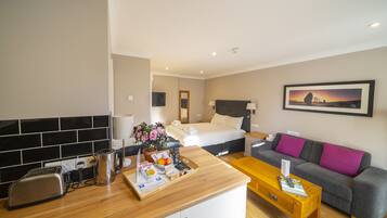 A hotel room with a kitchenette, a bed, a sofa, and a painting on the wall.