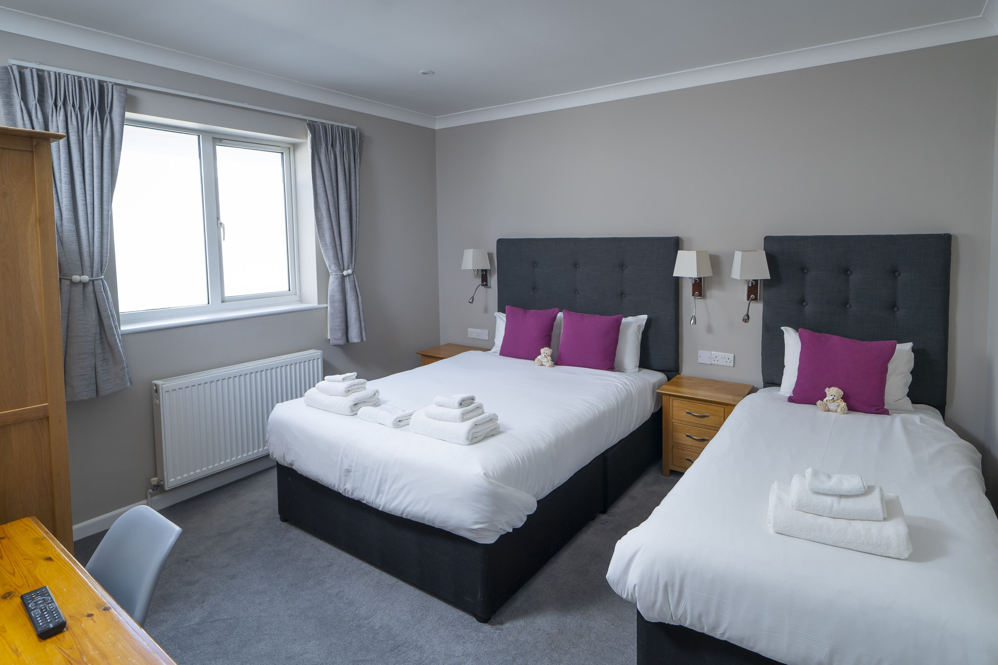 A hotel room with two beds, a wooden desk, a chair, a window with curtains, and a radiator.